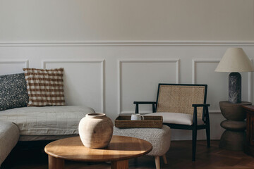 Serene cozy living room bathed in natural light. Neutral color palette. Wooden coffee table, wicker chair, tray with cup of tea coffee. Soft cushions on sofa, minimalistic decor. Empty wall bacground. © tabitazn
