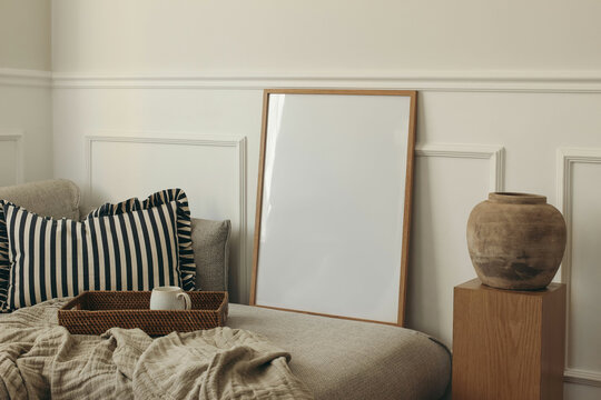 Empty wooden picture frame on sofa, pillow. Wicker tray with cup of tea, coffee. White panelled wall. Minimal elegant poster mockup. Artistic template. Wooden pedestal clay vase. Scandinavian interior