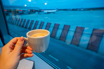 Hand holding coffee cup with night view of airport and planes in background. Concept of late-night flights, travel rituals, and quiet moments before departure