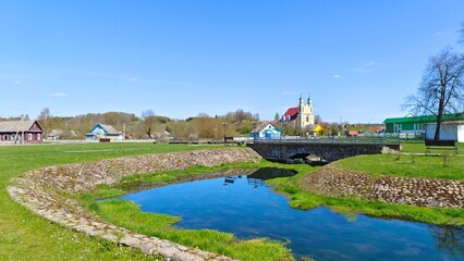 Obraz premium A small river flows among stone-lined grassy banks, which are connected by a stone footbridge with metal railings. Nearby are village houses and a church on a hill. Sunny spring weather and blue sky