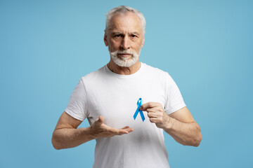 Senior man showing blue ribbon for prostate cancer awareness month isolated on blue background