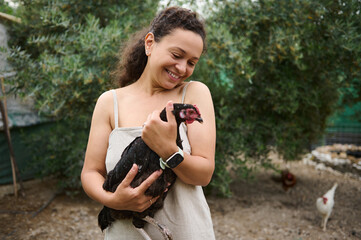 Smiling happy woman farmer holding and stroking free range hen, standing in a barn while feeding chickens and cockerels in the eco-farm. Eco farming and sustainable lifestyle concept. Animal love © Taras Grebinets