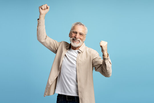 Euphoric senior man celebrating success with raised fists on blue background