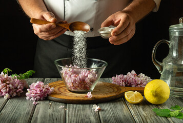 A chef skillfully adds sugar to a bowl of fresh flowers and prepares a lemon at a wooden counter in a cozy kitchen, showcasing meticulous culinary artistry. Making kvass with acacia