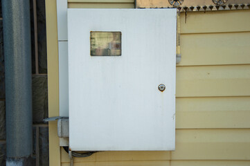A white utility box is mounted on the side of a yellow building, reflecting natural light in an urban setting during the day