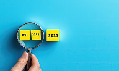 A hand holds a magnifying glass over two yellow blocks, each with a year printed on them (2024 and 2025). The blocks rest on a bright blue surface