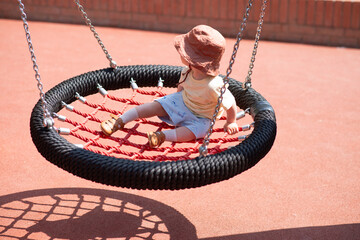 Happy toddler sitting on swing, dressed in summer cloves and wearing hat. Minimalistic background, modern outdoor playground. Activity for kids concept