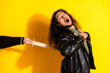 Young man in leather jacket pretending to be dramatically wounded with a wooden sword on yellow background