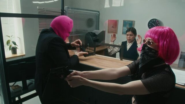 Nervous male robber masked with pink balaclava dropping handgun magazine with clumsy hands threatening teller with weapon while stealing money from bank with female partner