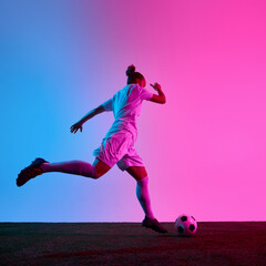 Female football player starting powerful kick toward ball during moment of action on field with neon gradient background. Concept of sport, impact, game, precision, motivation