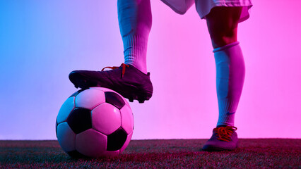 Detail view of female footballer's foot resting on ball on turf field under pink and blue lighting against gradient background. Concept of sport, control, readiness, visual power, brandimng.