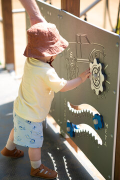 Baby boy dressed with summer cloves and hat is playing outside on the street, touching and exploring playground for little ones designed for sensory play. Lifestyle photo, sunny weather. 