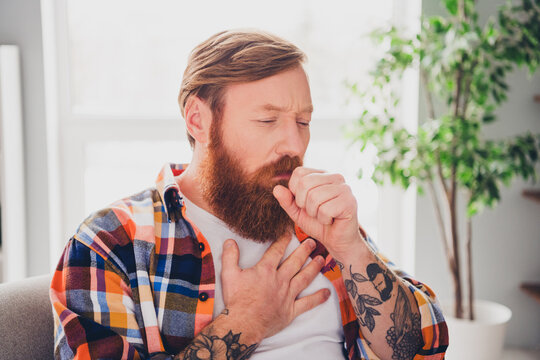 Bearded man in checkered shirt coughing while resting in a bright home interior with natural light and cozy decor