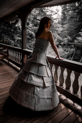 Elegant woman in a unique dress made of paper stands on a porch surrounded by nature