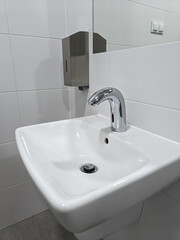 Close-up of a white ceramic sink with a modern automatic faucet and stainless steel soap dispenser mounted on a tiled bathroom wall.