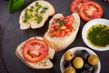Mediterranean snacks set. Olives, oil, herbs and sliced ciabatta on a wooden board on black slate stone board over dark background, juicy tomatoes on fresh bread, pesto as topping. top view. Flat lay
