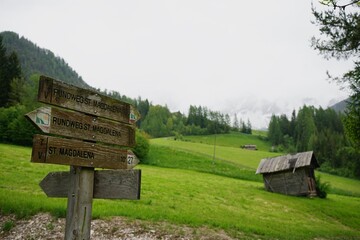 Editorial image. South Tyrol, Italy &ndash; May 22, 2024 &ndash; Wooden trail signs pointing toward St. Magdalena, with a backdrop of alpine meadows and misty mountains.