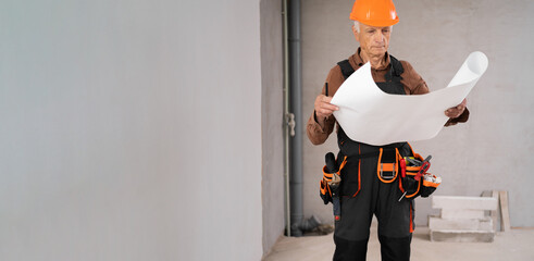 Elderly male builder studying layout plan new house, working with documents on construction site. Copy space