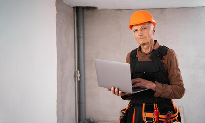 Elderly man in builder safety helmet looking at the camera and typing at laptop in home interior