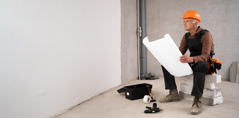 Senior man wearing hard hat at construction site. Construction worker reading house plan working in new building.