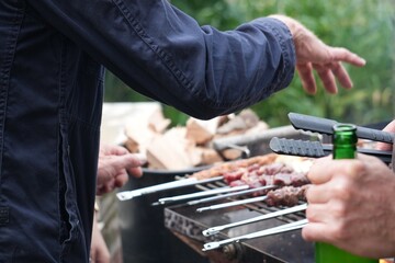Men grilling skewers on a charcoal barbecue while enjoying drinks and conversation, capturing a casual and social summer moment with focus on hands, food, and shared outdoor experience