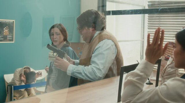 Shot from behind tellers counter of male unprofessional robber holding wads of money with excitement showing bundles of cash to relieved female partner while bank cashier standing with arms raised