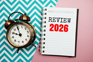 Review 2026 text on paper notebook with alarm clock top view on pink background