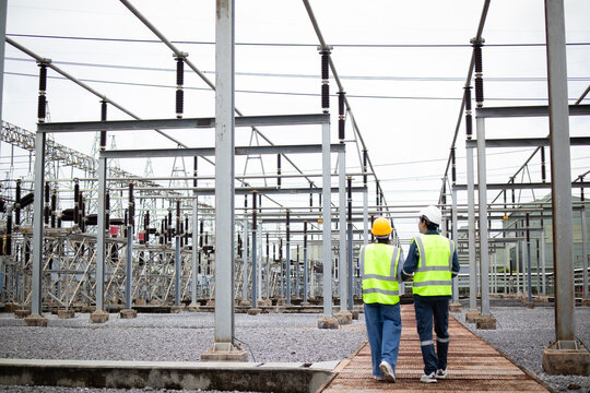 Engineer is working at high voltage substation wearing safety gear and reflective vest while inspecting electrical equipment in industrial outdoor environment