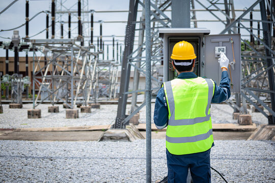 Engineer is working at high voltage substation wearing safety gear and yellow helmet while inspecting electrical equipment in industrial outdoor environment