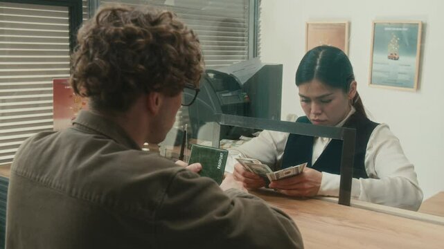 Female bank worker focused on counting dollar bills assisting male client with withdrawing funds from account then handling cash to customer at service window