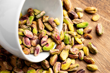 Shelled pistachio nuts spilled on wooden table from fallen white ceramic cup. Top view. Health food ingredient