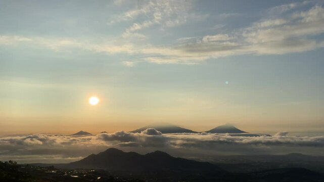 The view of the sunrise and slowly moving clouds from Mount Sumbing, Indonesia, with golden sunlight illuminating the sky and the magnificent silhouettes of Mount Merapi and Mount Merbabu.
