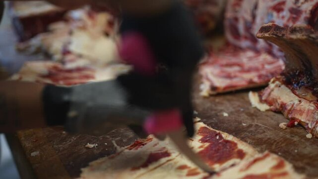 A butcher is cutting meat on a wooden table. The meat is raw and bloody. The butcher is wearing gloves and a watch