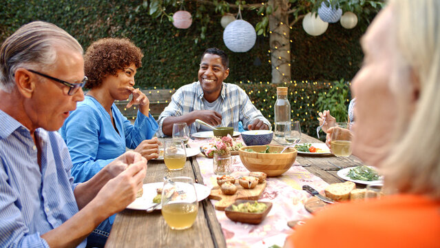 Group Of Mature Friends Sitting Around Table Enjoying Outdoor Meal In Backyard