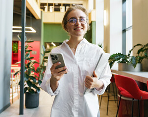 Bright Young Professional Smiles While Holding a Phone and Laptop in a Modern Office Filled With...