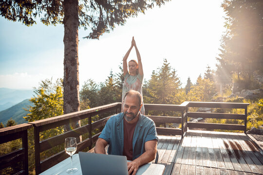 Man working remotely on laptop while woman does yoga outdoors with mountain view