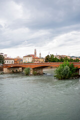 A scenic view of Verona along the Adige River, with historic buildings and lush hills in the background on a cloudy day