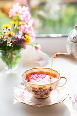 Creative postcard with porcelain cup of tea, teapot and a vibrant bouquet of garden flowers. Elegant tea set in beautiful morning sun light. Good morning and teatime concept. Slow life visual trend.