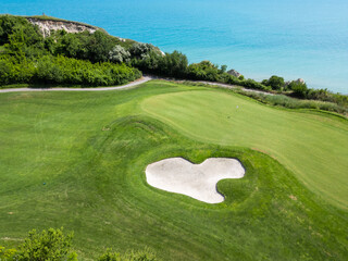 Scenic aerial view of a coastal golf course with green fairways, sand bunkers, golf carts, and players, surrounded by lush vegetation and turquoise sea