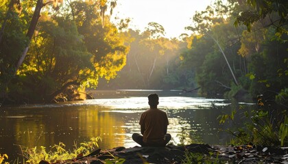 Person sits cross-legged at river, surrounded by sunlit green trees