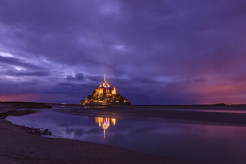 Mont Saint-Michel abbey illuminated at night, under stormy sky, with mirror reflection in calm water, listed as World Heritage by UNESCO, shortly after sunset, Normandy, Manche, France