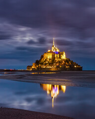 Mont Saint-Michel abbey illuminated at night, under stormy sky, with mirror reflection in calm water, listed as World Heritage by UNESCO, shortly after sunset, Normandy, Manche, France