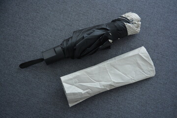 Things are objects of constant use. A beautiful beige, gray umbrella with a black lining and a cover lies folded on a gray fabric background.