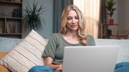 Fototapeta premium Young gorgeous woman typing on laptop sitting on sofa working online. Beautiful busy relaxed female using gadget to communicate. Charming blonde lady in living room. Working from home concept.
