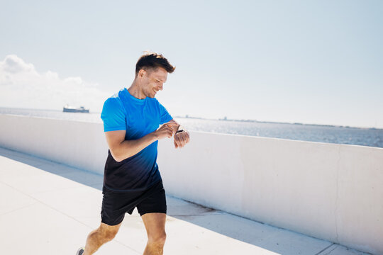 Energetic Jogger Checks Watch During Sunny Coastal Run, Feeling Invigorated by Ocean Breeze and Vibrant Sky