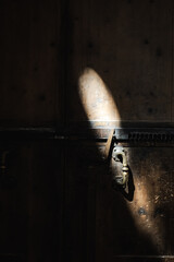 Narrow beam of sunlight illuminates antique door handle in dramatic shadow, creating moody and mysterious scene