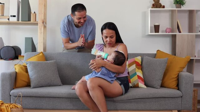 Loving hispanic mother bottle feeding her baby girl while sitting on a comfortable sofa in the living room, with the caring father standing next to them, creating a heartwarming family scene