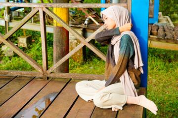 Asian Muslim woman sits cross-legged on the wooden floor, leaning against a pillar while massaging...