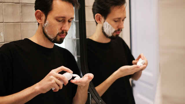 Young man applying shaving foam to cheeks while looking in mirror
