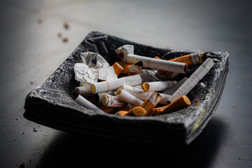 Close-up of a black ashtray filled with cigarette butts, ash, and paper trash, placed on a dark table with scattered debris.